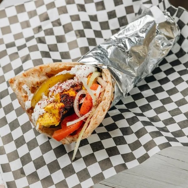 MidEast Eats chicken shawarma wrap with pickles, onions, tomatoes, and sauce on checkered paper.