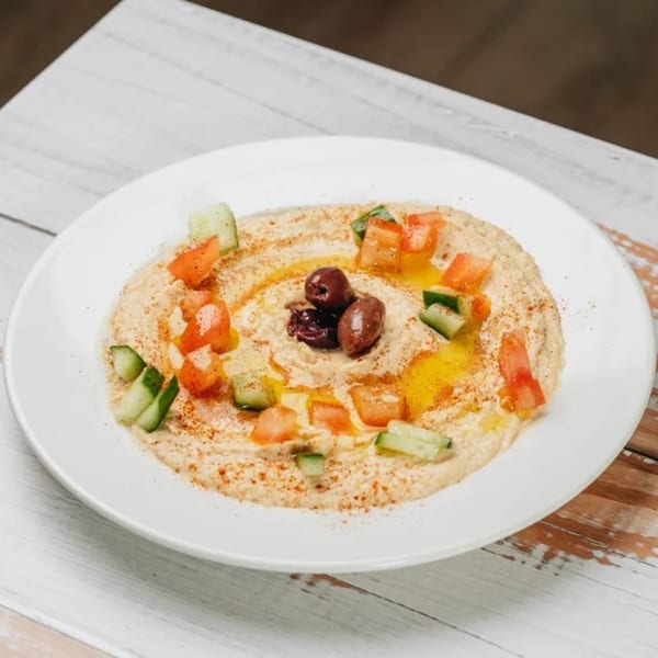 MidEast Eats hummus with olives, diced tomatoes, cucumbers, and olive oil on a white plate.