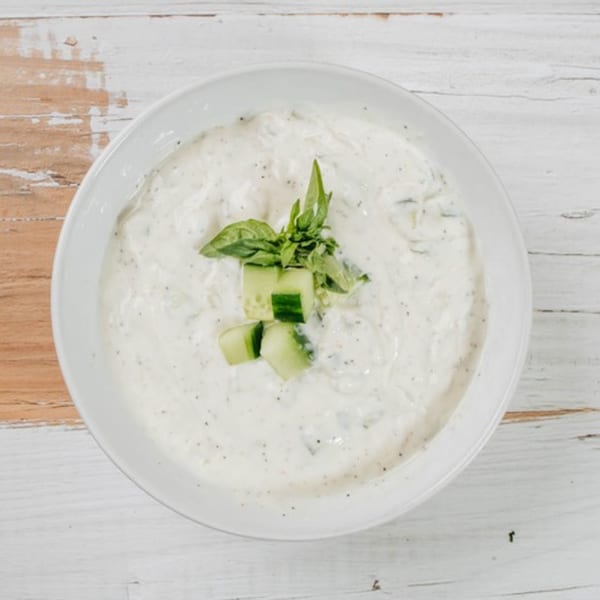 MidEast Eats' creamy tzatziki dip, garnished with fresh cucumber chunks and basil, on a white wooden table.