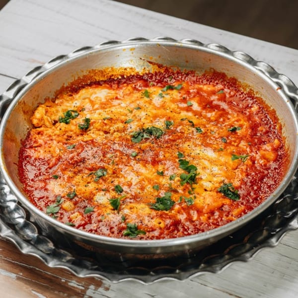A close-up of a Middle Eastern dish, possibly shakshuka, in a metal pan with a red sauce and white cheese.